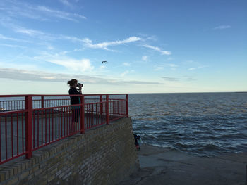 Side view of woman photographing on pier by sea against sky