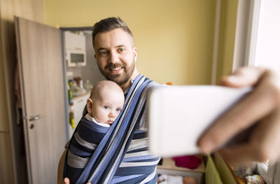 Father with baby son in sling at home taking a selfie