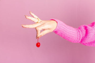 Female hand wearing pink sweater and holding a cherry. light pink background.