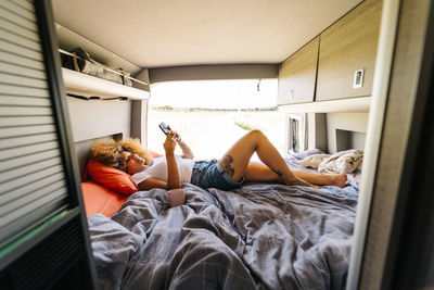 Anonymous woman with afro hairstyle lying upside upon bed in trailer and browsing mobile phone on sunny summer day