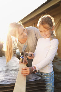 Happy mother looking at daughter hammering nail on plank outside house being renovated