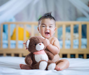 Portrait of shirtless baby boy holding stuffed toy while sitting on bed