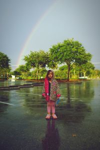 Portrait of girl standing on road against rainbow during monsoon