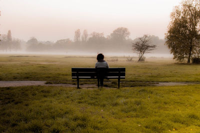 Rear view of woman sitting on bench at park during foggy weather