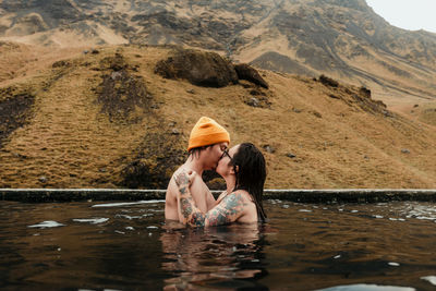 Side view of young tattooed hipsters kissing and hugging in water between mountains