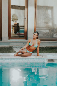 Portrait of young woman in bikini sitting at poolside against house