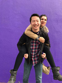 Smiling young man piggybacking girlfriend against purple wall