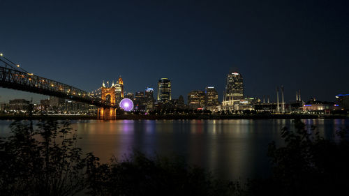 Illuminated bridge over river by buildings against sky at night