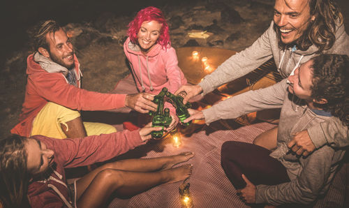 High angle view of friends toasting beer bottles while sitting on blanket at night
