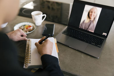 Hands of businessman writing in diary while doing video call with female colleague on laptop
