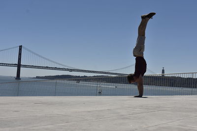 Side view of man doing handstand on footpath by river against clear sky