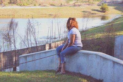 Full length of woman looking at lake while sitting on retaining wall