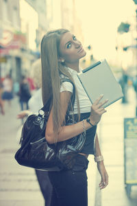 Portrait of young woman holding digital tablet while standing on city street