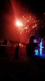 Boy holding illuminated laser sword while sitting on field against firework display at night