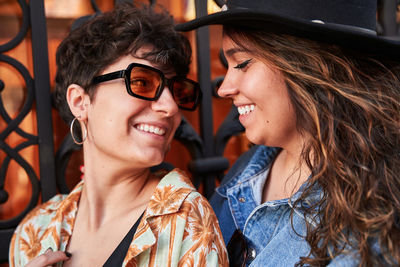 Happy young women in stylish clothes smiling and looking at each other against metal gate during romantic date on street of madrid, spain