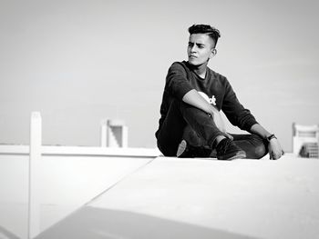 Young man sitting on retaining wall against clear sky