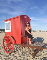 Portrait of woman wearing sunglasses sitting on cart at beach against sky