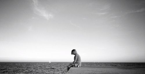 Side view of woman sitting on retaining wall by sea against sky