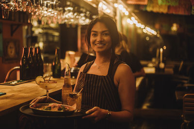 Portrait of smiling waitress holding food and drinks on serving tray while working at bar