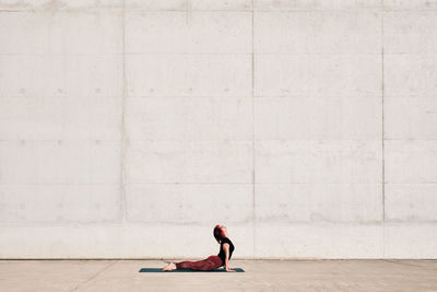 Side view of trendy fit female athlete in sportswear doing baby cobra yoga exercise on sports mat while training alone on street against concrete wall in sunny day