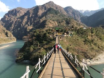 People walking on footbridge over river against mountains