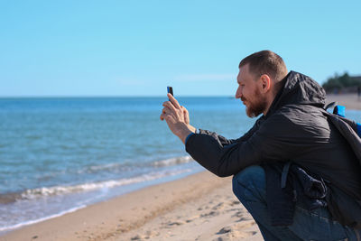 Bearded man in black raincoat on blue sea landscape background with smartphone in hands takes selfie