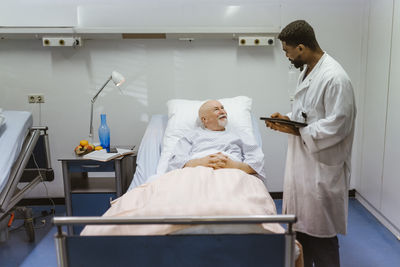 Young male doctor with digital tablet talking to senior patient lying on bed at hospital