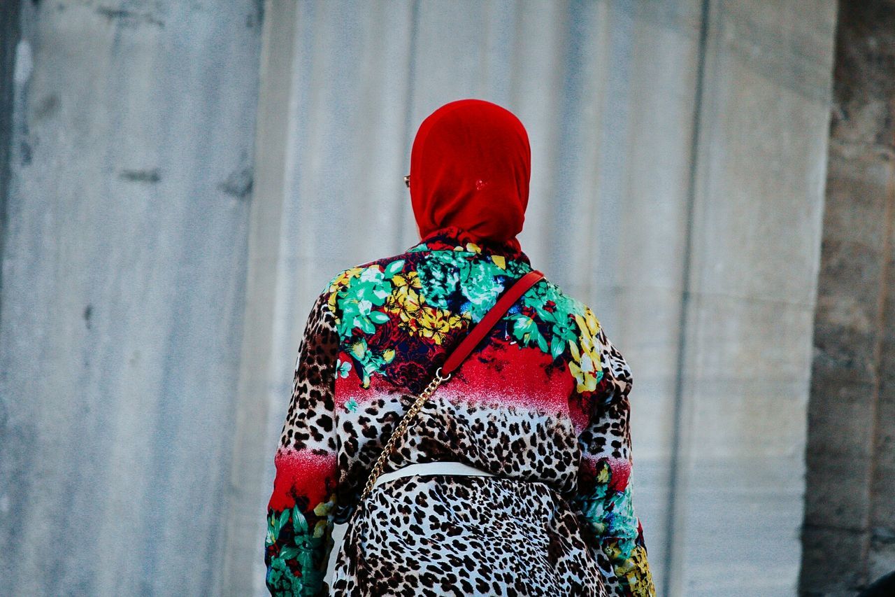 rear view, real people, clothing, one person, day, pattern, focus on foreground, lifestyles, red, standing, wall - building feature, leisure activity, winter, adult, women, outdoors, hat, casual clothing, multi colored, warm clothing, scarf, floral pattern, hairstyle