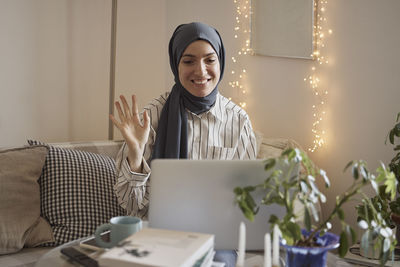 Happy female freelancer waving on video call over laptop while working in living room at home