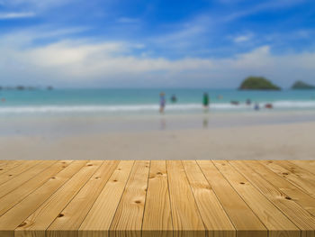 Dark brown plank wooden board with blurred relaxing peoples on the beach with cloudy blue sky.