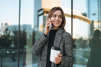 Young smiling woman in coat with coffee cup using mobile phone in evening city street