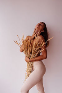 Young shirtless woman holding plants standing by wall