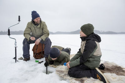 Smiling man talking with boy while son doing ice fishing at frozen lake