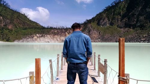 Rear view of man standing footbridge over lake against mountains