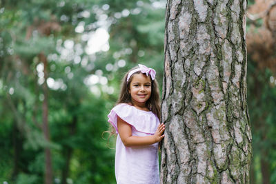 A beautiful girl in a pink dress stands near a tree in the forest in summer, smiling and dreaming