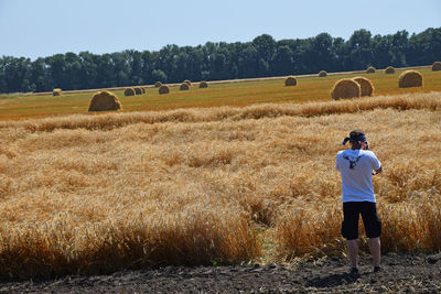 Rear view full length of man photographing hay bales by wheat field