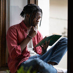 Concentrated inquisitive african american man student reading book sits on windowsill near window