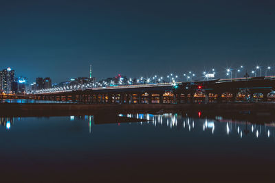 Illuminated bridge over river by buildings against sky at night