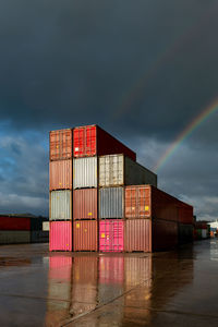 A stack of shipping containers on a deserted commercial dock with sunlight and rainbow