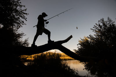 Silhouette man holding fishing rod standing by lake against sky during sunset