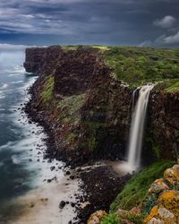 Scenic view of waterfall by sea against cloudy sky