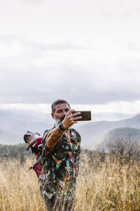 Man with dog in backpack taking photos on the mountain with cloudy sky