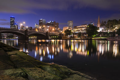 Illuminated bridge over river by buildings against sky at night