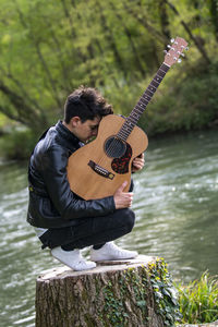 Young man playing guitar while sitting by lake