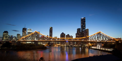 Illuminated bridge over river by buildings against sky in city