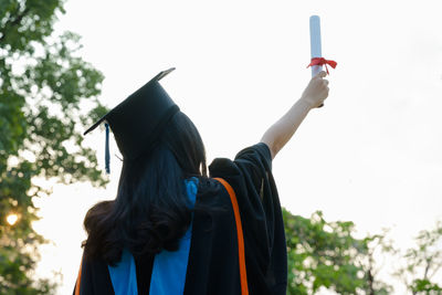 Rear view of woman in graduation gown holding diploma against sky