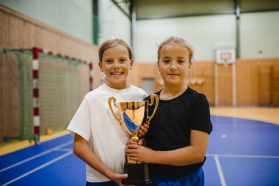 Portrait of smiling girls holding trophy while standing together in sports court
