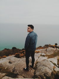 Man standing on rock looking at sea shore against sky