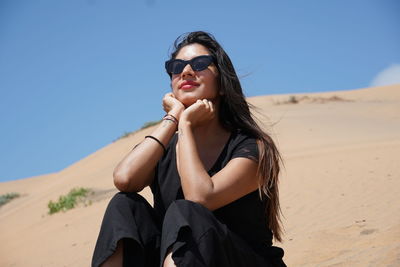 Young woman wearing sunglasses on sand at desert against clear blue sky