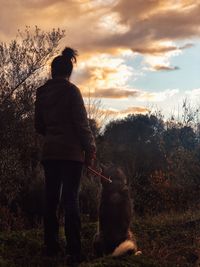 Rear view of woman with dog standing against sky during sunset
